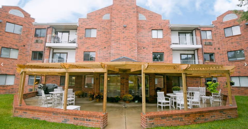 Outdoor patio area with a wooden pergola attached to a red brick building. The patio has white chairs and tables arranged for seating, with some potted plants and a grill visible. The building has multiple windows and balconies.