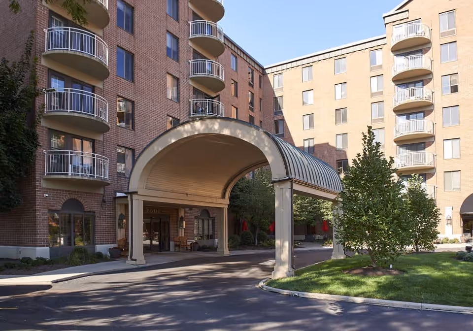 Exterior view of a multi-story senior living facility with brick walls and several balconies. The entrance features a large covered driveway with an arched roof, surrounded by landscaped greenery and trees under a clear blue sky.