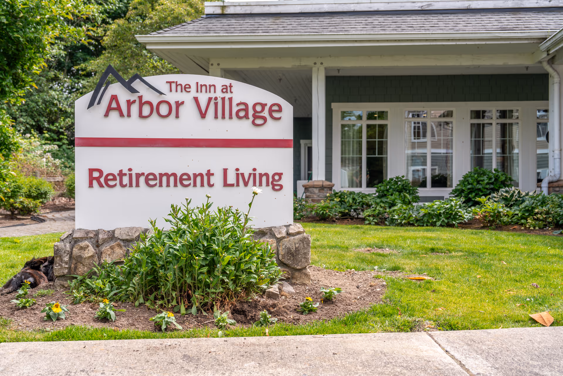 Outdoor view of the entrance area of Arbor Village Senior Living showing a sign that reads 'The Inn at Arbor Village Retirement Living' surrounded by greenery and flowers with part of the building visible in the background.