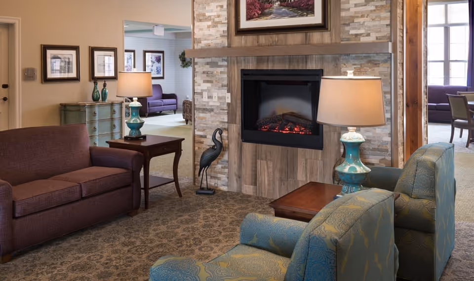 A cozy living room area with a stone and wood fireplace in the center. There are two blue patterned armchairs and a brown sofa arranged around the fireplace. Two wooden side tables with turquoise lamps are placed beside the chairs and sofa. In the background, there are additional seating areas with purple sofas and framed artwork on the walls.