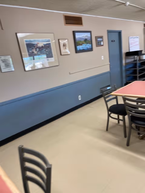 Interior room with beige and blue walls, several framed pictures hanging on the wall, a door, and a table with chairs around it. The floor is light-colored tile.