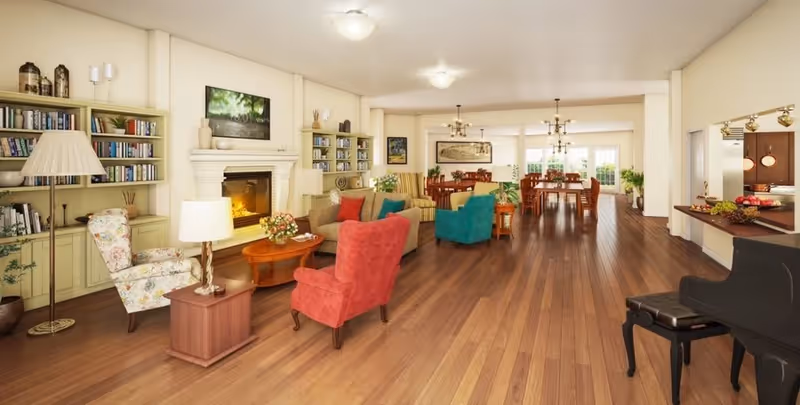 A spacious and well-lit living room area with wooden flooring, featuring a fireplace surrounded by built-in bookshelves filled with books and decorative items. The room has multiple seating options including a floral armchair, a red armchair, a teal armchair, and a beige sofa with colorful cushions. There are two wooden coffee tables with flower arrangements. In the background, there is a dining area with wooden tables and chairs, and large windows letting in natural light. A black grand piano is visible on the right side near a pass-through window to the kitchen.