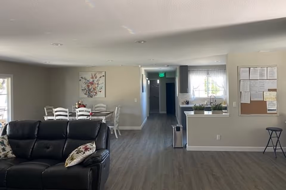 Interior view of a senior living facility common area with a black leather sofa in the foreground, a dining table with white chairs and a floral centerpiece in the background, a kitchen area with a window and sink, and a bulletin board on the wall. The floor is wood-style laminate and the walls are painted light gray.
