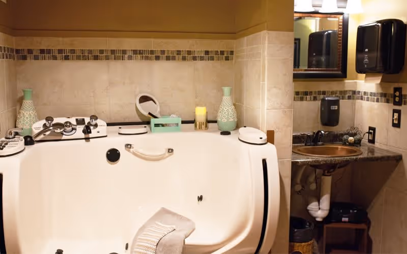 A bathroom featuring a large white walk-in bathtub with a towel draped over the edge. The walls are tiled with a decorative border near the top. On the bathtub ledge, there are two green decorative vases, a candle, and a small mirror. To the right, there is a sink with a dark countertop, a mirror above it, and a black soap dispenser mounted on the wall.