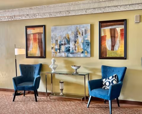 A cozy seating area with two blue upholstered armchairs, one with a patterned pillow, positioned on either side of a glass console table. The table holds decorative items including a white bust sculpture and a silver bowl. Behind the table, three framed abstract paintings hang on a light yellow wall. A floor lamp stands next to the left armchair, and the floor is covered with a patterned carpet.