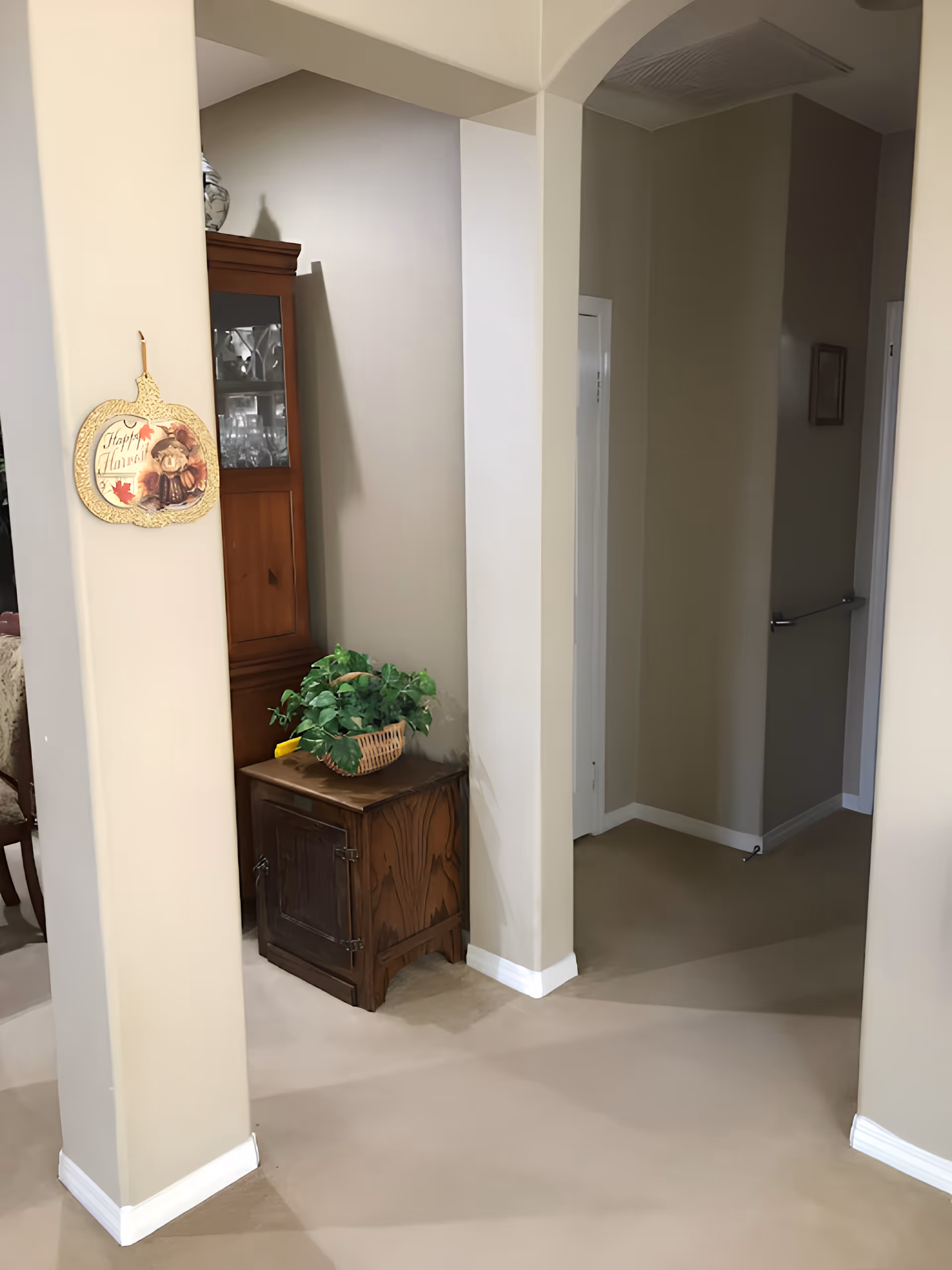 Interior view of a hallway area with beige walls and carpeted floor. A wooden cabinet with glass doors is positioned against the wall, topped with a basket containing a green leafy plant. A decorative hanging on a column reads 'Happy Harvest' with an image of a bear and autumn leaves. The hallway leads to other rooms with visible doorways and a handrail on the right wall.