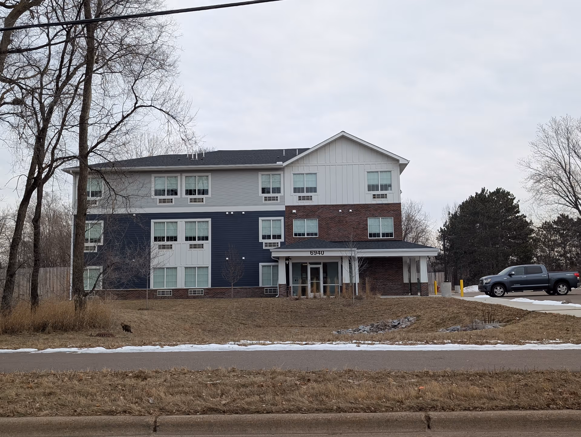 Exterior view of a three-story building with a mix of brick and siding facade, several windows, and a covered entrance with the number 6940 above it. There are leafless trees and a parked pickup truck on the right side, with patches of snow on the ground and a cloudy sky.