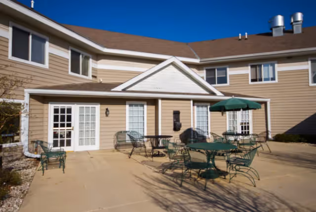 Outdoor patio area of a senior living facility with several green metal tables and chairs, one table with a green umbrella, beige siding building with white-framed windows and doors under a clear blue sky.