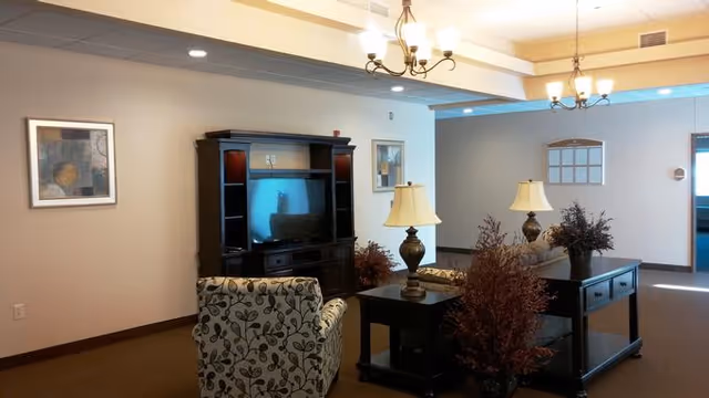A cozy living room area in an assisted living facility featuring a floral patterned armchair, a dark wood entertainment center with a TV, two table lamps on side tables, decorative plants, and framed artwork on beige walls under ceiling lights.