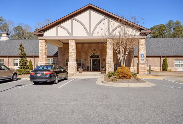 Front exterior view of Oaks at Shiloh Point facility showing a covered entrance with stone pillars, a brick facade, parked cars, and some landscaping with trees and shrubs.