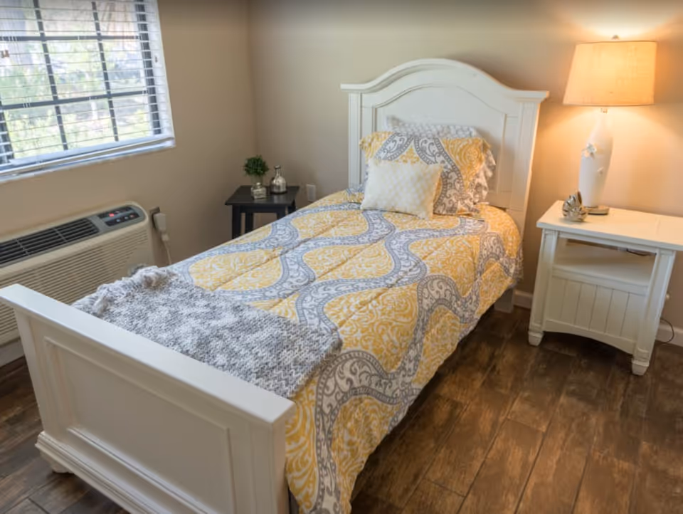 A cozy bedroom with a single white wooden bed featuring a yellow and gray patterned bedspread and matching pillows. There is a gray knitted throw blanket at the foot of the bed. To the right of the bed is a white nightstand with a white lamp and a decorative item. To the left, there is a small black side table with a small plant and a decorative bottle. A window with blinds and an air conditioning unit is on the left wall. The floor is wooden.