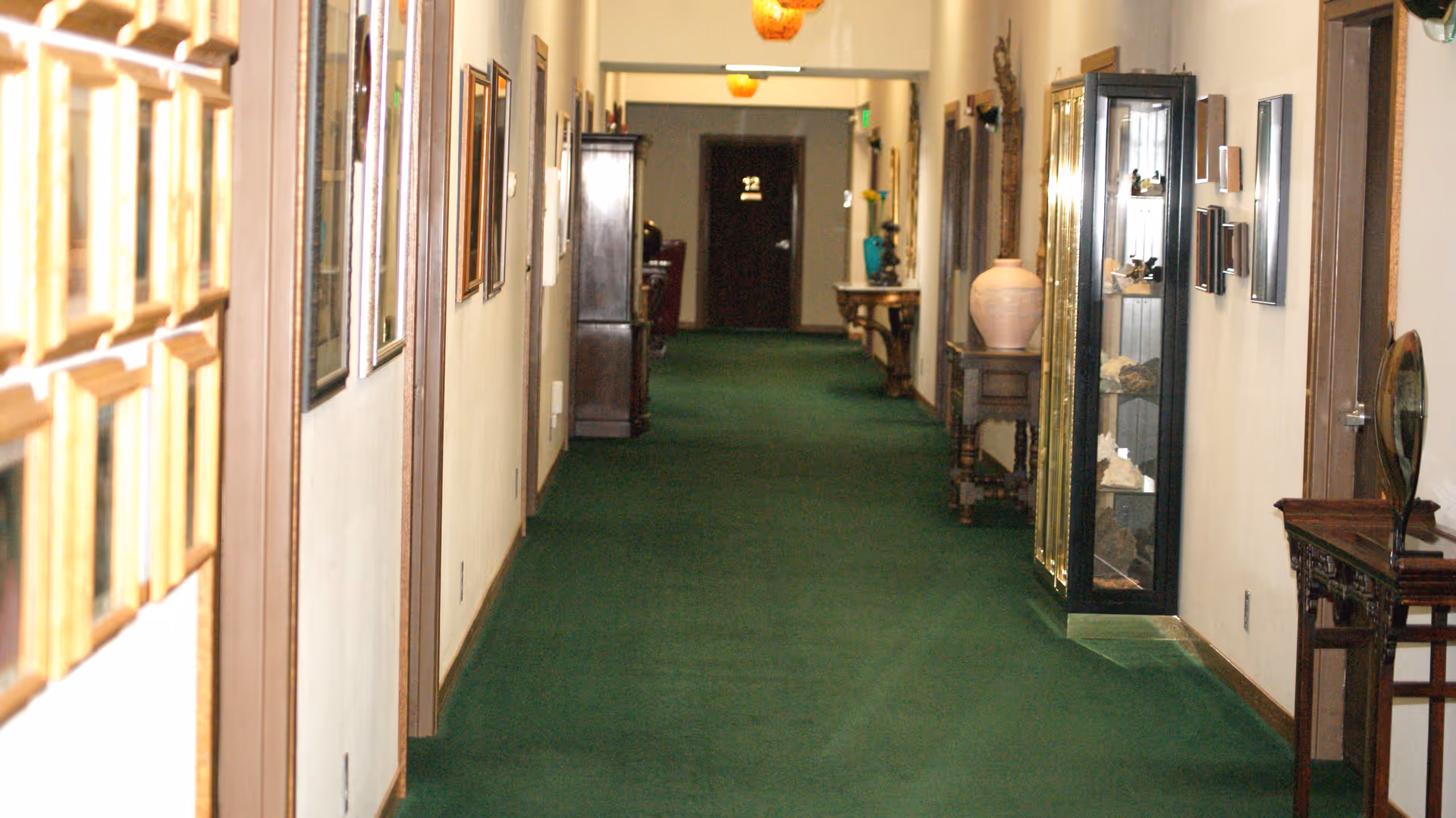 Long carpeted hallway with green carpet, framed artwork on the walls, display cabinets and small tables along the sides.