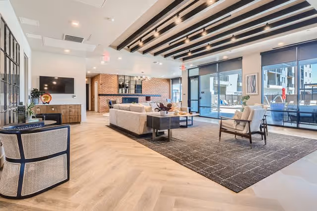 A spacious and modern living room area with light wood flooring, a large dark area rug, a beige sectional sofa, a wooden coffee table, and a cushioned armchair. The room features large windows with roller shades, allowing natural light to fill the space. There is a flat-screen TV mounted on the wall above a wooden console, and a brick accent wall with a bar area and stools in the background. The ceiling has exposed beams with recessed lighting.