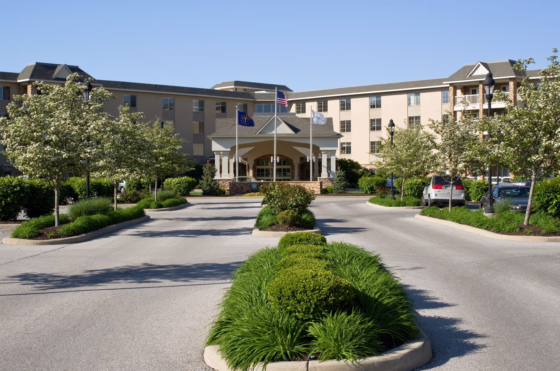 Front exterior view of Westside Village Nursing Center showing a multi-story building with a covered entrance, three flagpoles with flags, landscaped greenery, and a driveway with parked cars on a clear sunny day.