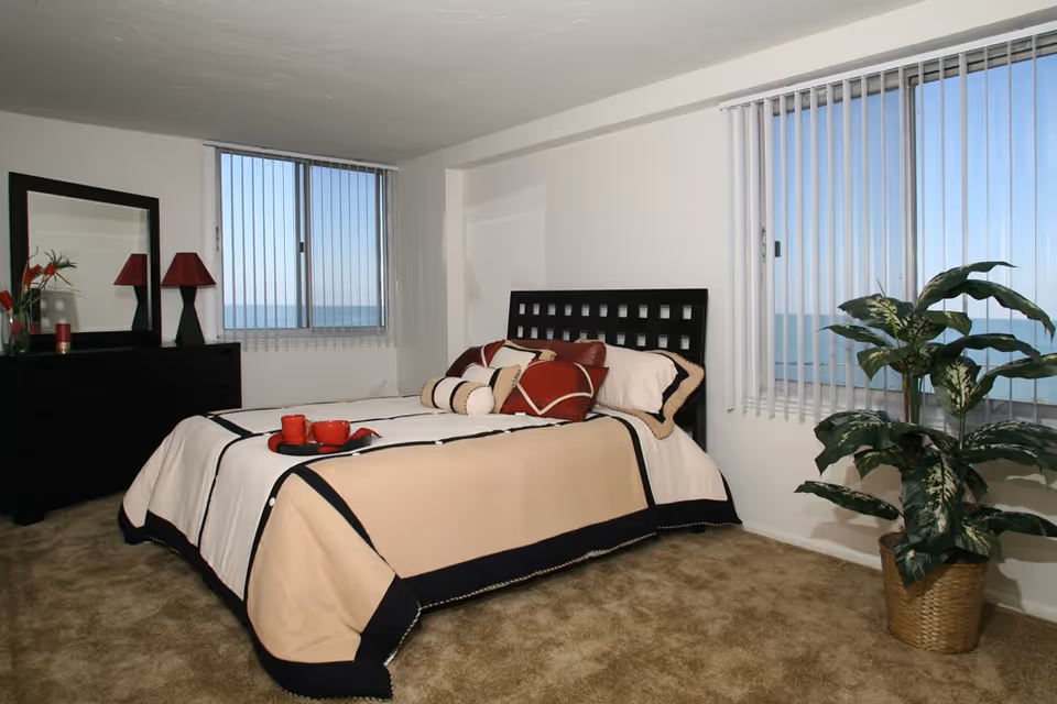 A bedroom with a bed featuring beige, black, and white bedding with red accent pillows. There is a black dresser with a mirror and a red lamp on the left side of the room. Two windows with vertical blinds show a view of the ocean. A large green potted plant is placed near the window on the right side. The floor is carpeted in a light brown color.
