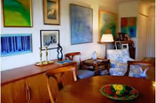 Interior room with a round dining table in the foreground, wooden sideboard and chairs, upholstered armchairs, and colorful paintings on the walls.