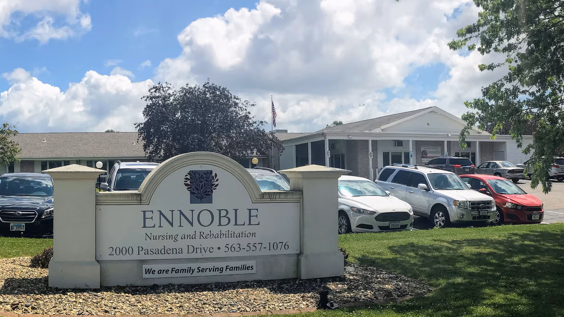 Exterior view of Ennoble Nursing and Rehabilitation facility with a sign in the foreground displaying the name, address, and phone number. Several cars are parked in front of the building under a partly cloudy sky.