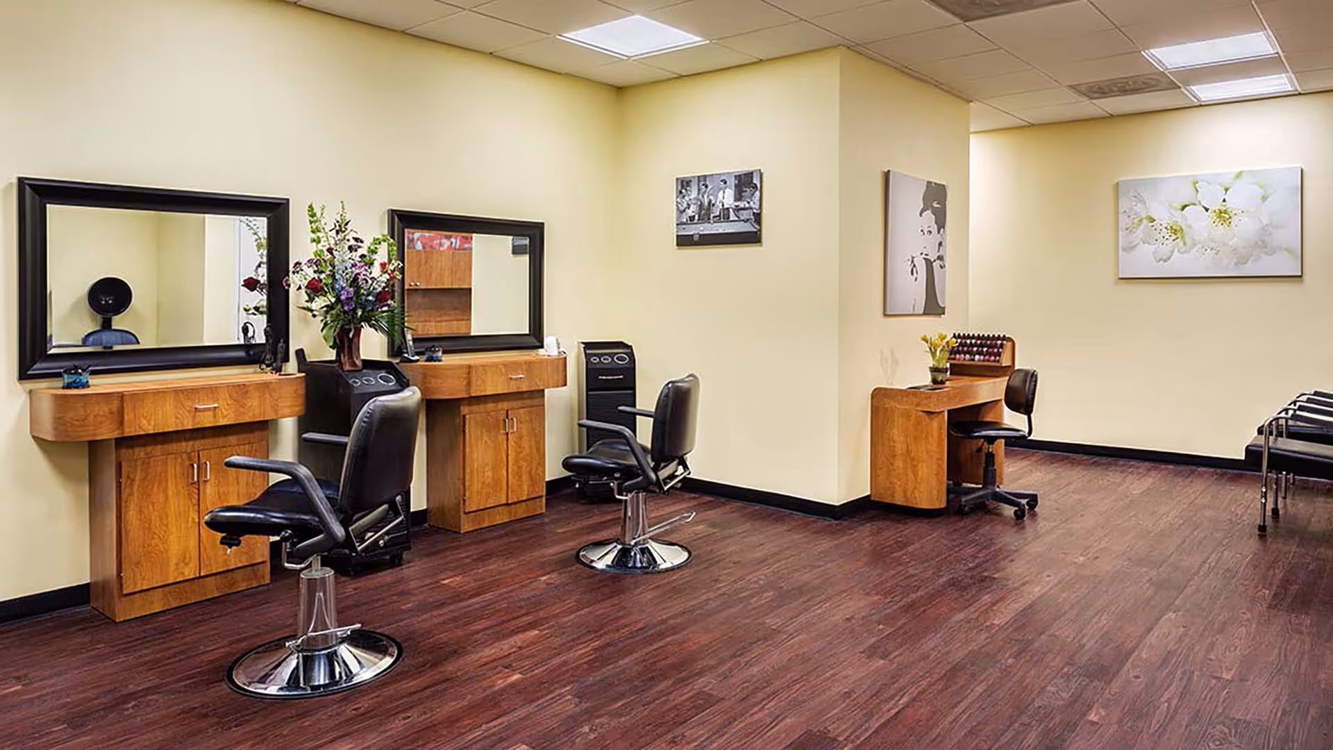 Interior view of a salon area with two styling stations featuring large mirrors, wooden cabinets, and black salon chairs. There is a small desk with a chair and nail polish display on the right side, along with framed artwork on the walls and dark wood flooring.