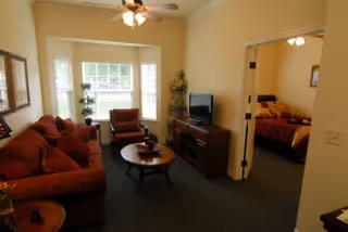 Furnished living room with sofa and chairs facing a TV cabinet, bay window, and an open doorway to a bedroom.