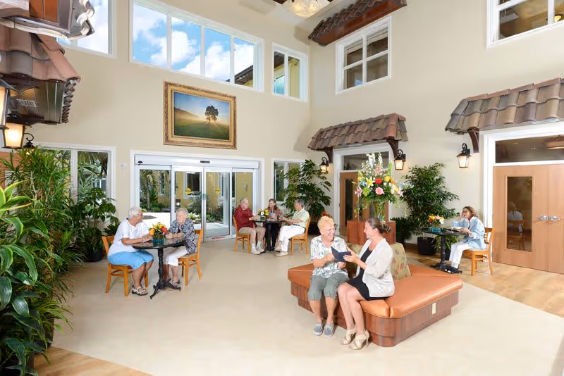 A bright, spacious common area in a senior living facility with high ceilings and large windows. Several elderly residents are seated at small tables and on a cushioned bench, engaging in conversation and activities. The room is decorated with plants, wall art, and warm lighting fixtures, creating a welcoming atmosphere.