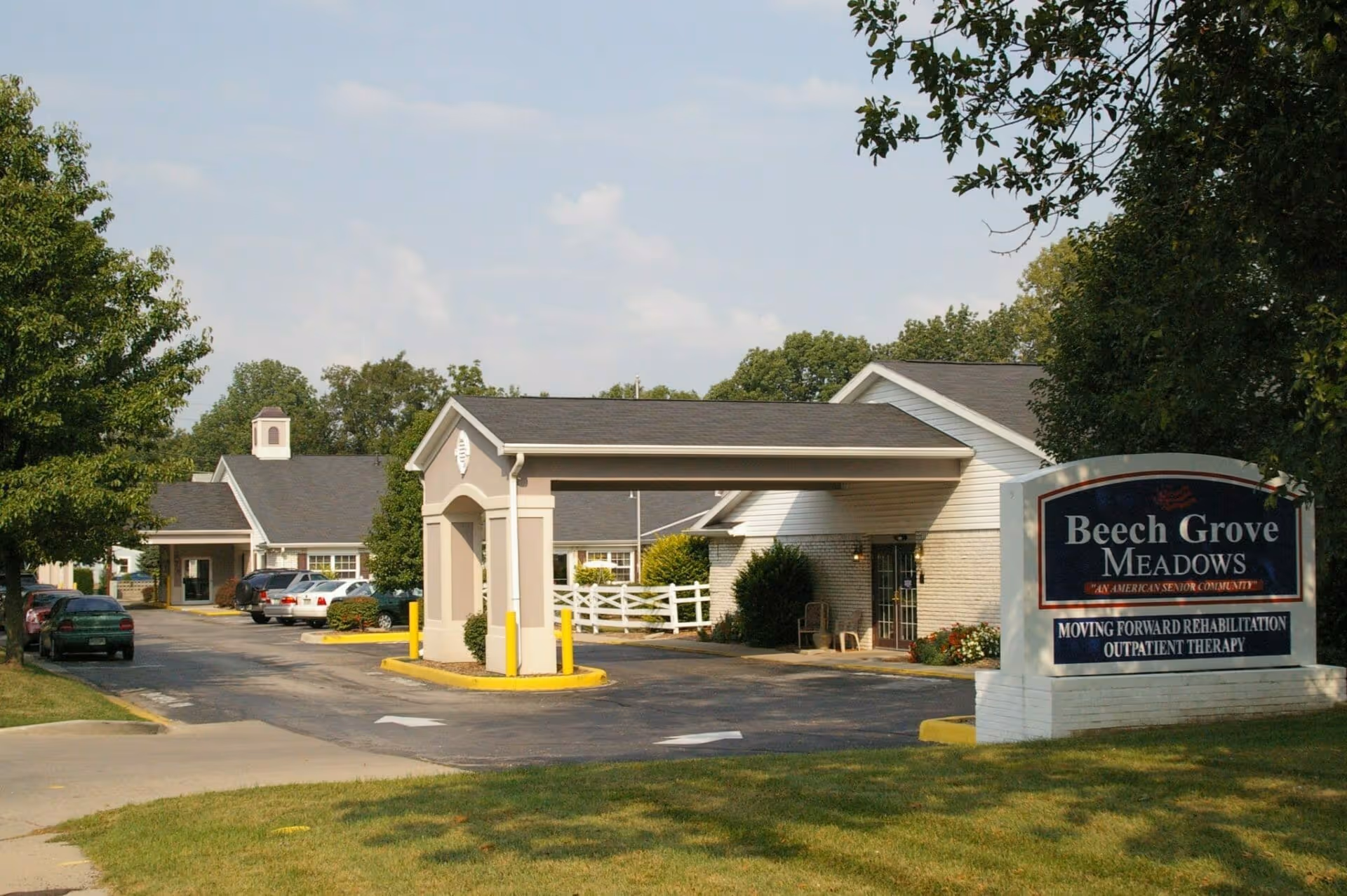 Exterior view of Beech Grove Meadows senior living facility showing the entrance with a covered drop-off area, several parked cars, trees, and a large sign with the facility name and services offered.