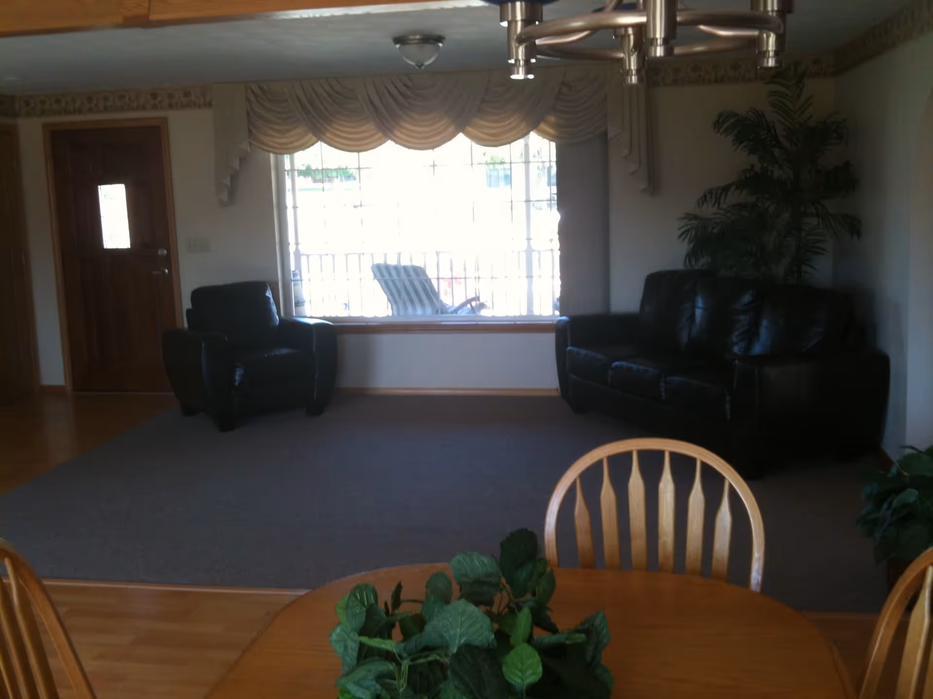 Interior view of a living room area with a large window covered by draped curtains, a black leather armchair and sofa, a wooden door, a ceiling light fixture, and a wooden dining table with chairs and a green plant centerpiece.