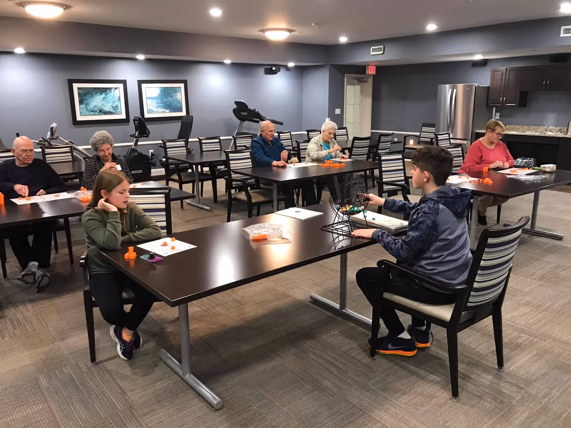 A group of elderly people and two children sitting at tables in a spacious room playing a game with orange markers and bingo cards. The room has exercise equipment in the background, framed artwork on the walls, and a kitchenette area with a refrigerator and cabinets.