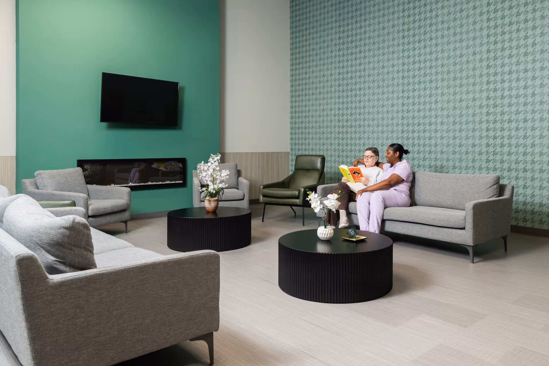 A modern living room with gray sofas and armchairs arranged around two black round coffee tables with flower vases. A flat-screen TV is mounted on a green accent wall above a sleek fireplace. Two women, one elderly and one caregiver in purple scrubs, sit on a sofa reading a book together. The room has light-colored flooring and a patterned green wallpaper on one wall.
