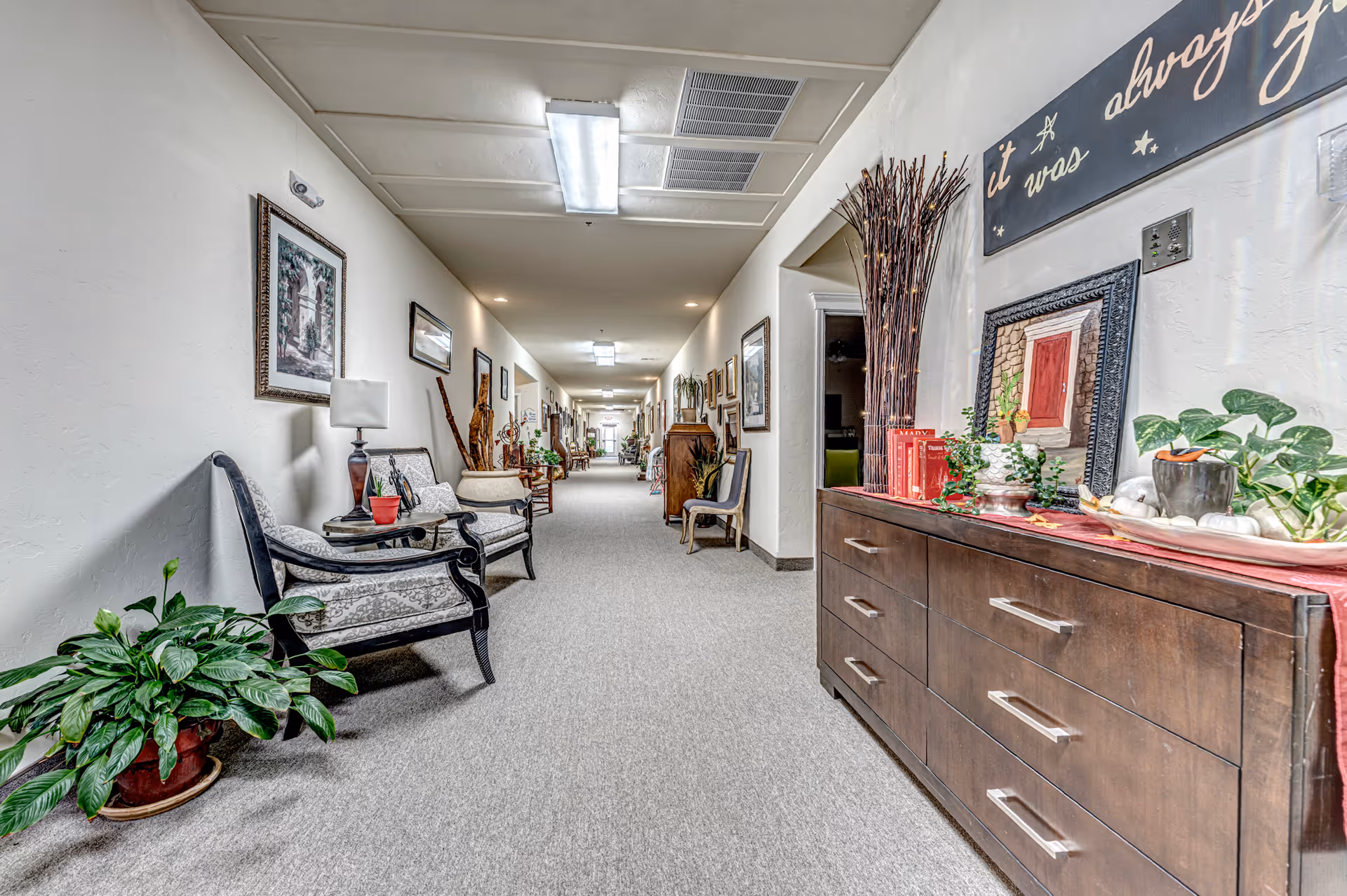 A long, well-lit hallway in a senior living facility with carpeted floors and white walls. The hallway is decorated with framed pictures, plants, and various pieces of furniture including chairs, a wooden dresser with decorative items, and a lamp. The ceiling has fluorescent lighting and ventilation panels.