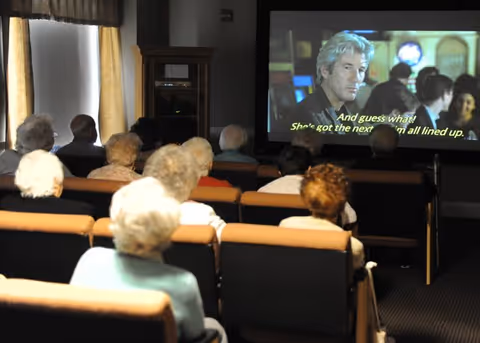 A group of elderly people seated in rows of chairs watching a movie projected on a screen in a dimly lit room with curtains on the windows.