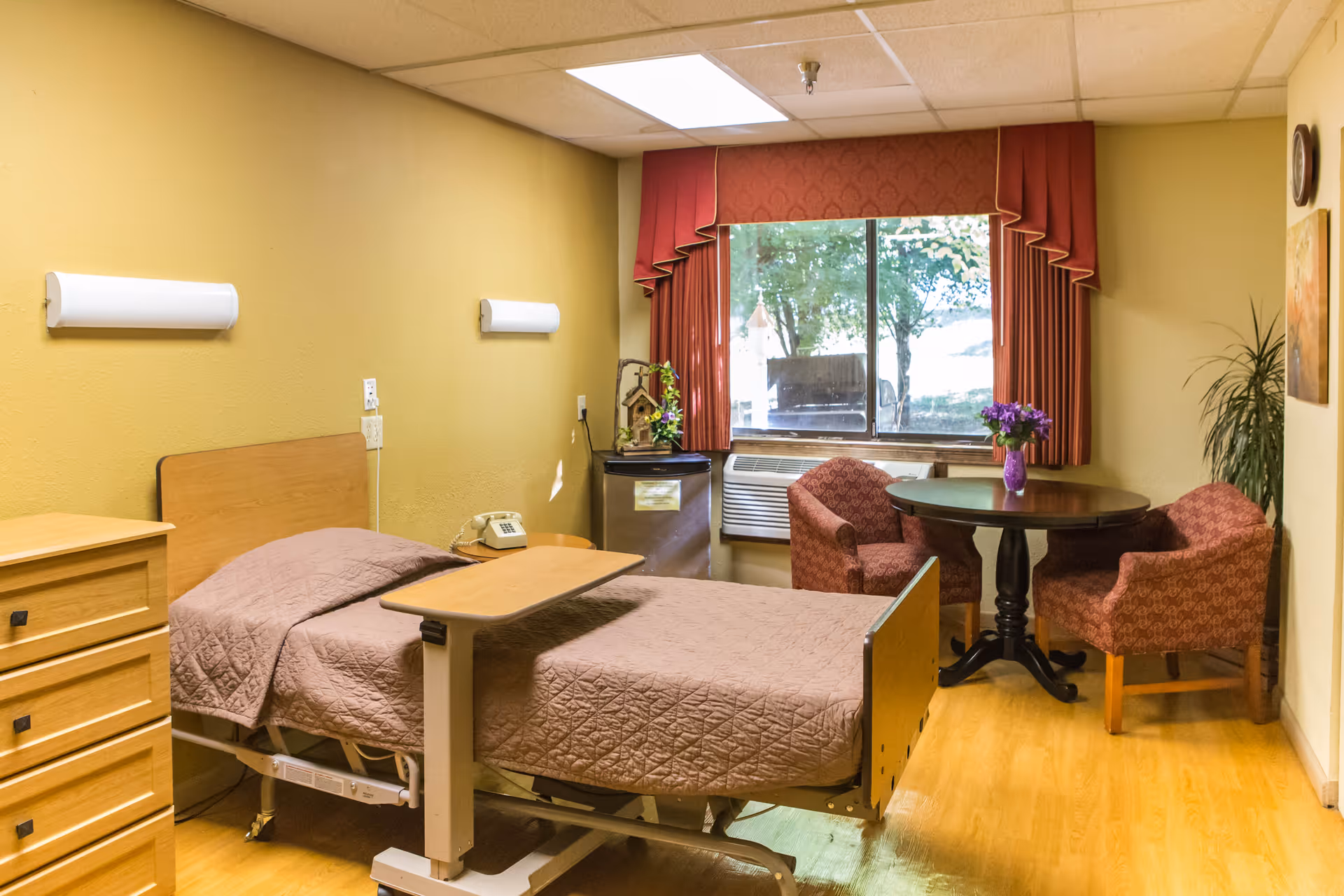 A senior living facility room with a hospital-style bed covered in a brown quilted blanket, a wooden dresser, a small table with a telephone, and a round table with two upholstered chairs near a window with red curtains. There is a small refrigerator and an air conditioning unit below the window, with a plant and wall decorations in the room.