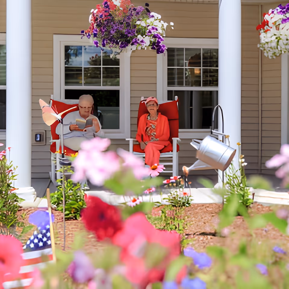 Two elderly women sitting on red cushioned rocking chairs on a porch of a beige building with white columns. There are colorful flowers in the foreground and hanging flower baskets above them. One woman is reading a book while the other is looking ahead. A watering can is hanging on the right side of the porch.