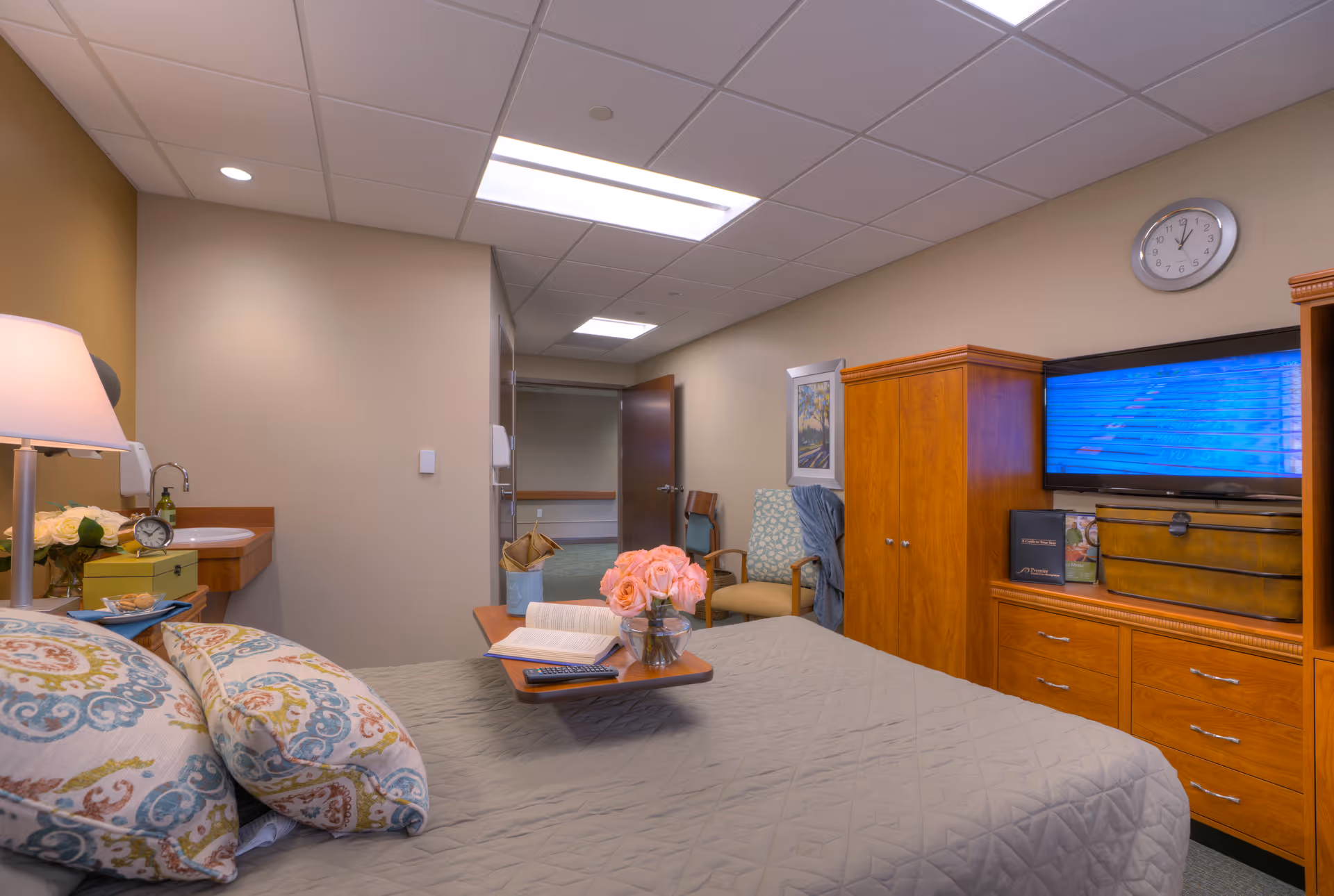 A cozy bedroom in a senior living facility featuring a bed with patterned pillows and a gray quilt. A small table on the bed holds a vase of pink flowers, an open book, and a remote control. The room includes wooden furniture such as a dresser with a TV mounted above it, a wardrobe, and a chair with a blanket draped over it. A clock is mounted on the wall above the TV, and a small sink with a lamp and decorative items is visible to the left. The door to the room is open, showing a hallway outside.