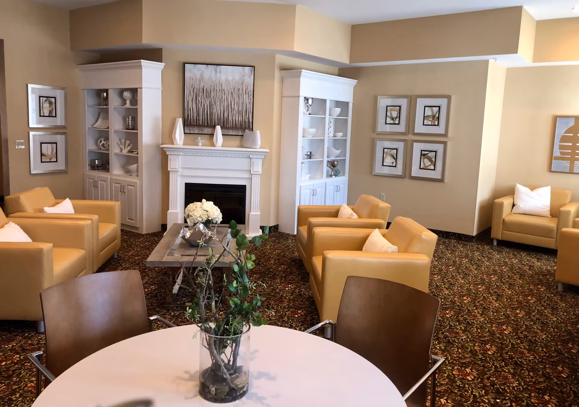 A cozy living room area with beige walls and patterned carpet. The room features four tan leather armchairs arranged around a wooden coffee table with a floral centerpiece. There is a white fireplace with decorative vases on the mantel and built-in white shelving units on either side containing various decorative items. The walls are adorned with framed abstract artwork. In the foreground, there is a round white table with two brown chairs and a glass vase with branches and greenery.