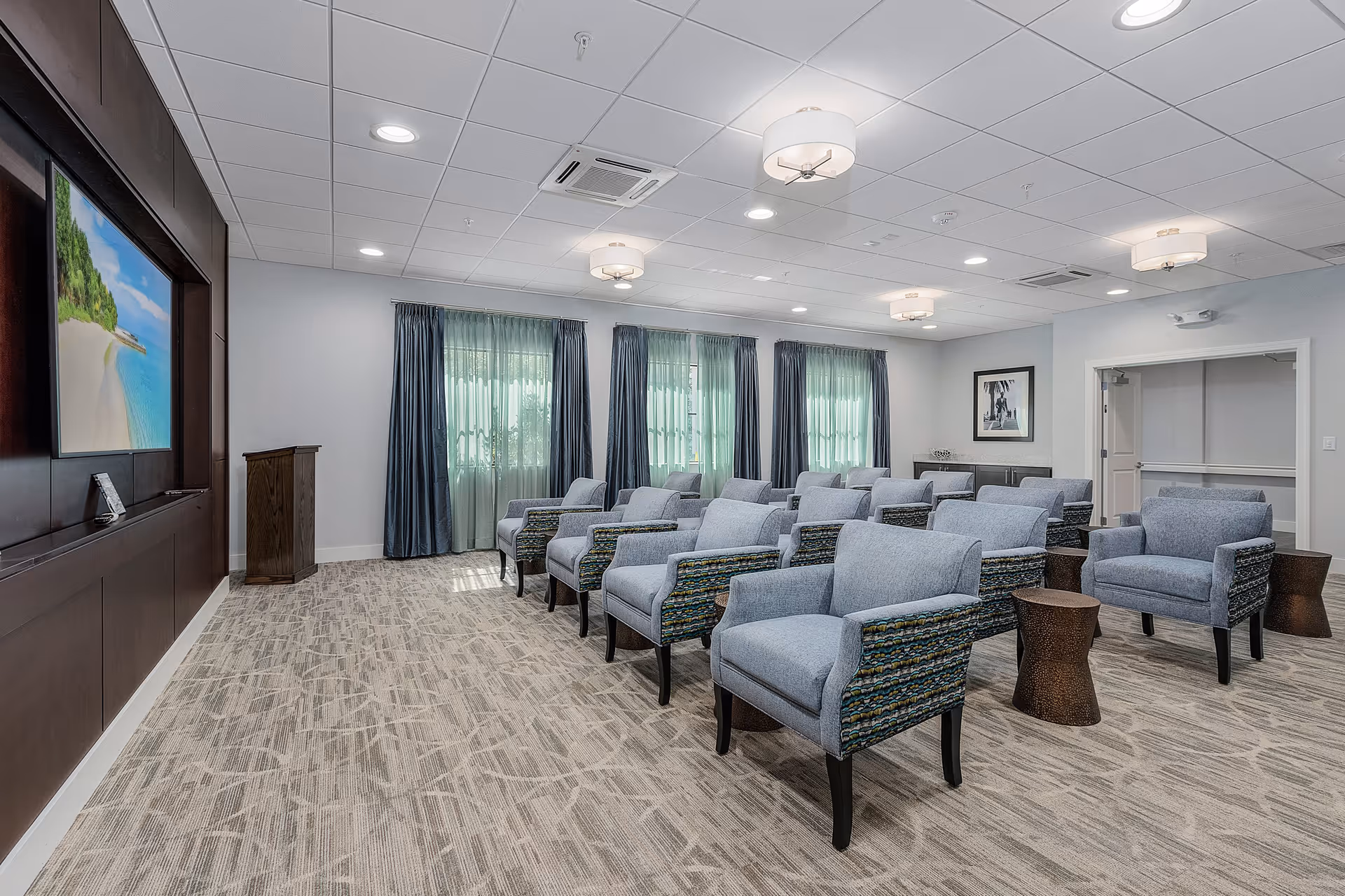 A spacious room with multiple rows of comfortable armchairs facing a large wall-mounted screen displaying a beach scene. The room has carpeted flooring, three large windows with blue curtains, and modern ceiling lights. There are small side tables next to some chairs and a wooden podium near the screen.