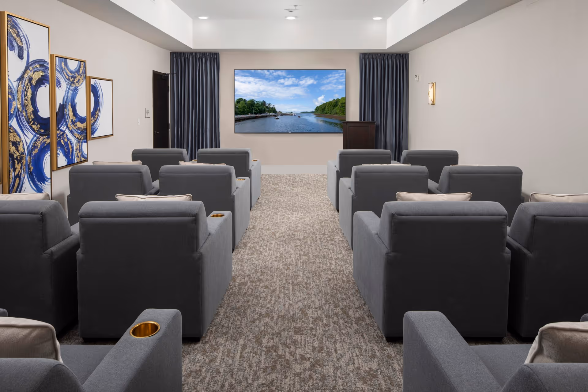 Carpeted screening room with rows of gray upholstered recliner chairs facing a wall-mounted television.