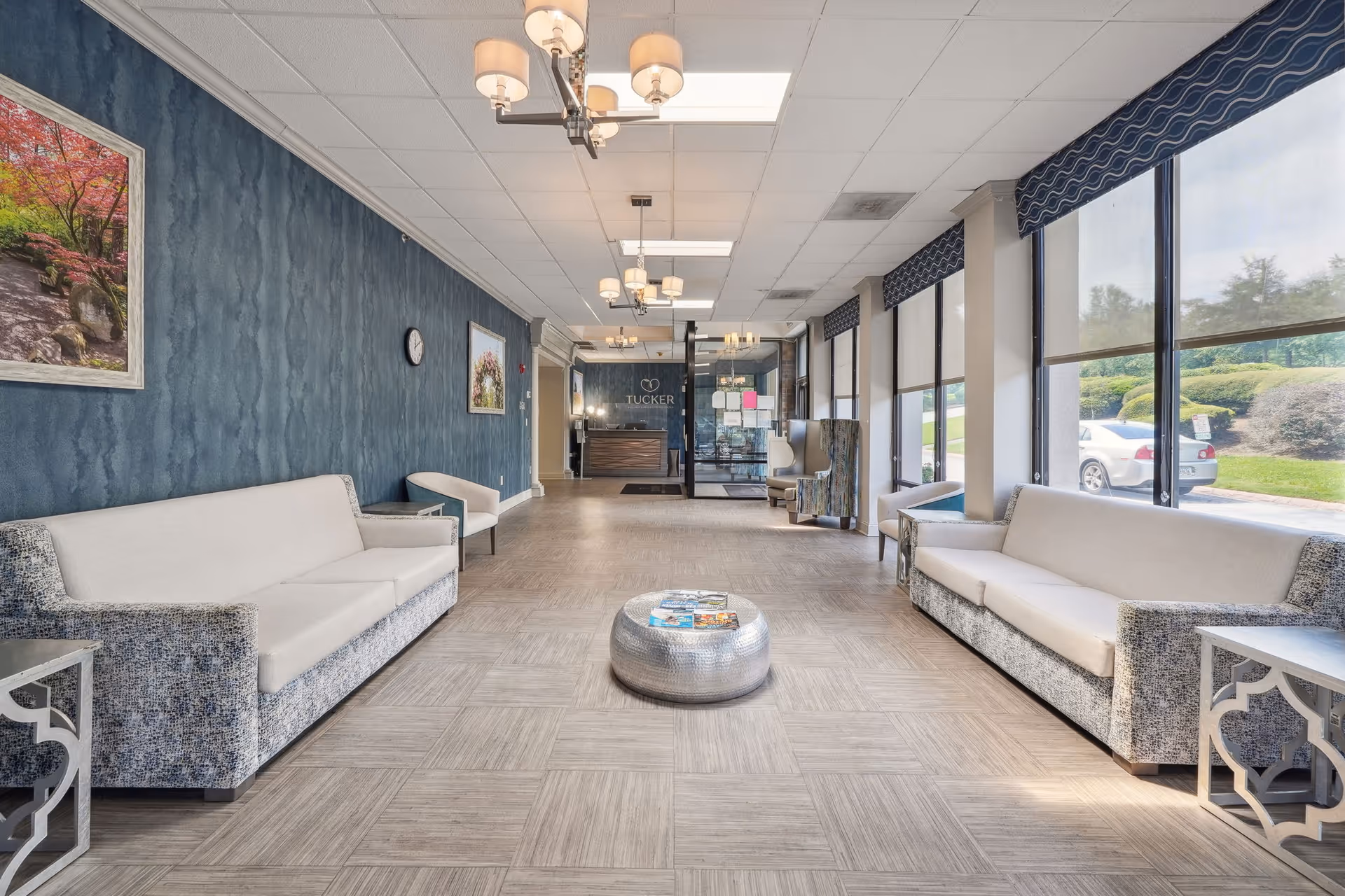 A spacious and well-lit waiting area or lobby with two long sofas facing each other, a round metallic coffee table with magazines, large windows with roller shades on the right side, blue textured walls with framed pictures, and a reception desk at the far end with the name 'Tucker' visible behind it.