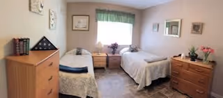 A small, neatly arranged bedroom with two single beds separated by a wooden nightstand. Each bed has a light-colored bedspread. The room features two wooden dressers, one on each side of the beds, with decorative items including flowers and framed pictures on the walls. A window with a green valance allows natural light into the room.