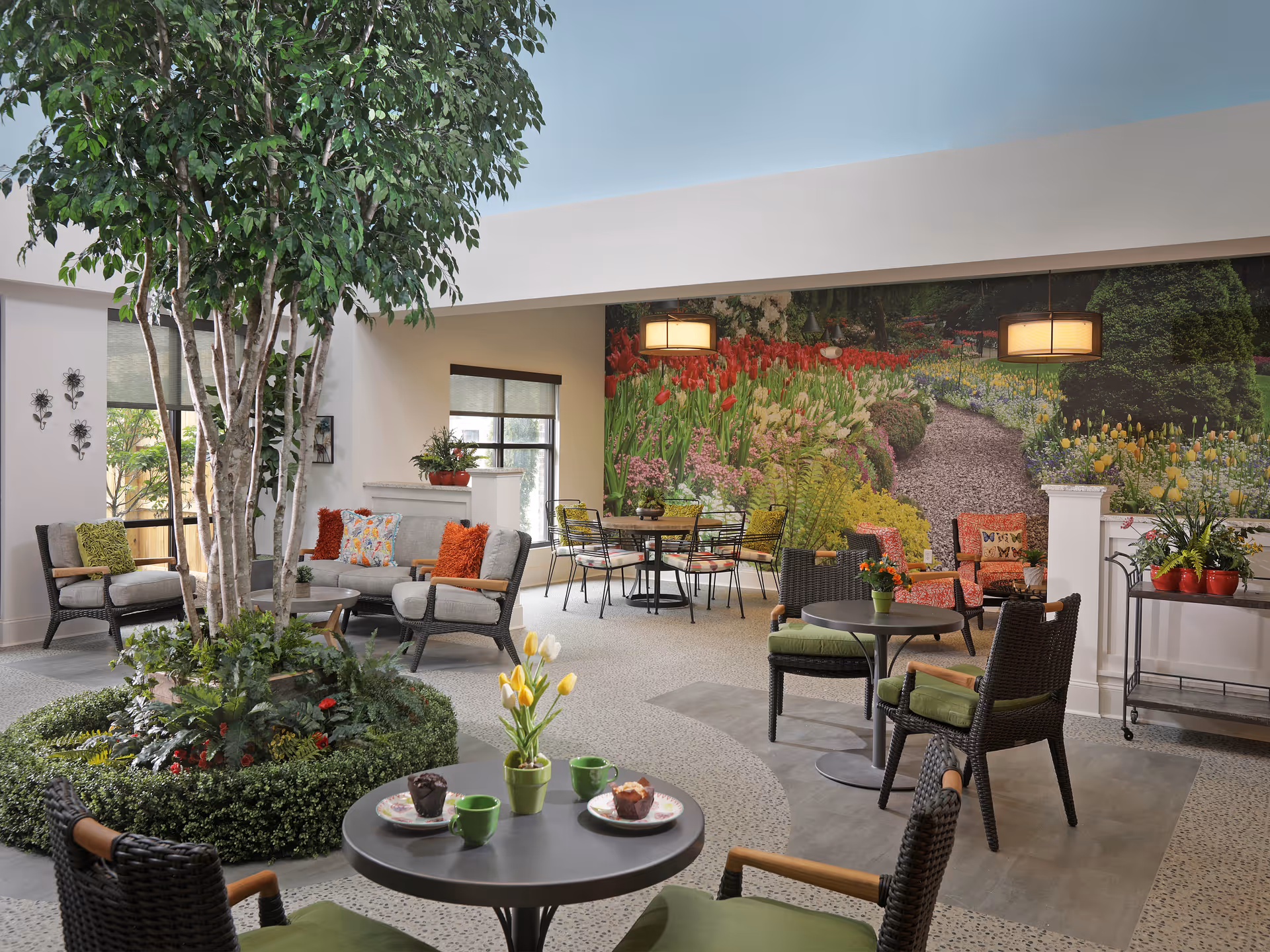 Bright communal lounge with wicker seating, round tables, an indoor tree planter, and a large floral mural on the back wall.