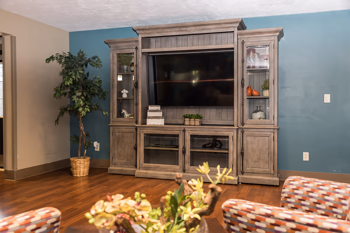 A living room area with a large wooden entertainment center holding a flat-screen TV. The entertainment center has glass cabinets displaying decorative items. To the left is a potted artificial tree, and in the foreground, there are patterned armchairs and a table with a floral arrangement. The walls are painted in beige and teal colors, and the floor is wooden.