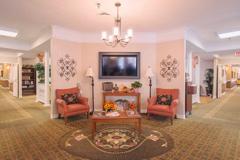 A cozy sitting area in a senior living facility with two red armchairs, a wooden coffee table with magazines, a wooden console table with decorative items and plants, a wall-mounted flat screen TV, two floor lamps, and decorative wall art. The area is carpeted with a patterned rug and is flanked by two hallways with bookshelves and plants.