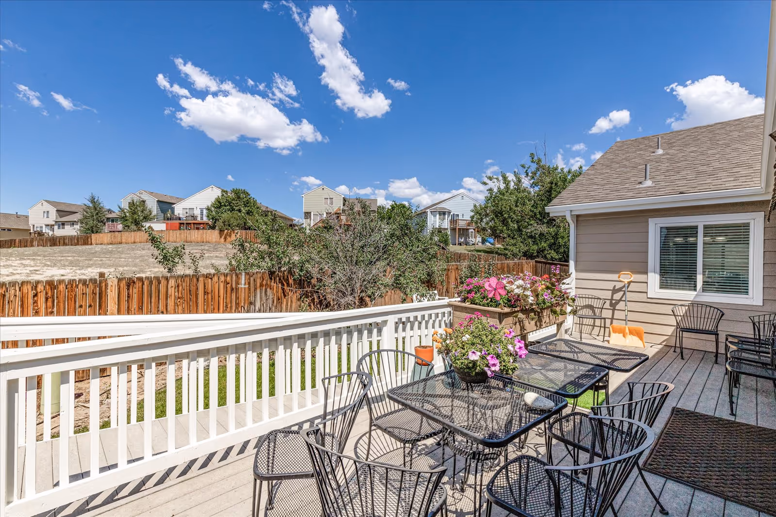 Outdoor patio area with black metal tables and chairs, flower pots with colorful flowers on the tables and railing, a beige building with a window, and a wooden fence enclosing the area. The sky is blue with scattered white clouds.