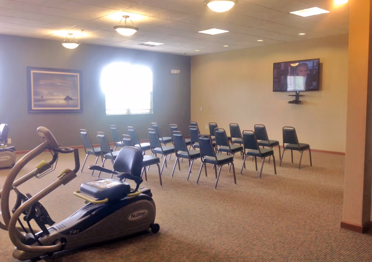A room with exercise equipment including a NuStep recumbent cross trainer in the foreground and several rows of chairs arranged facing a wall-mounted television. The room has beige walls, carpeted floor, ceiling lights, a window with blinds, and a framed picture on the wall.