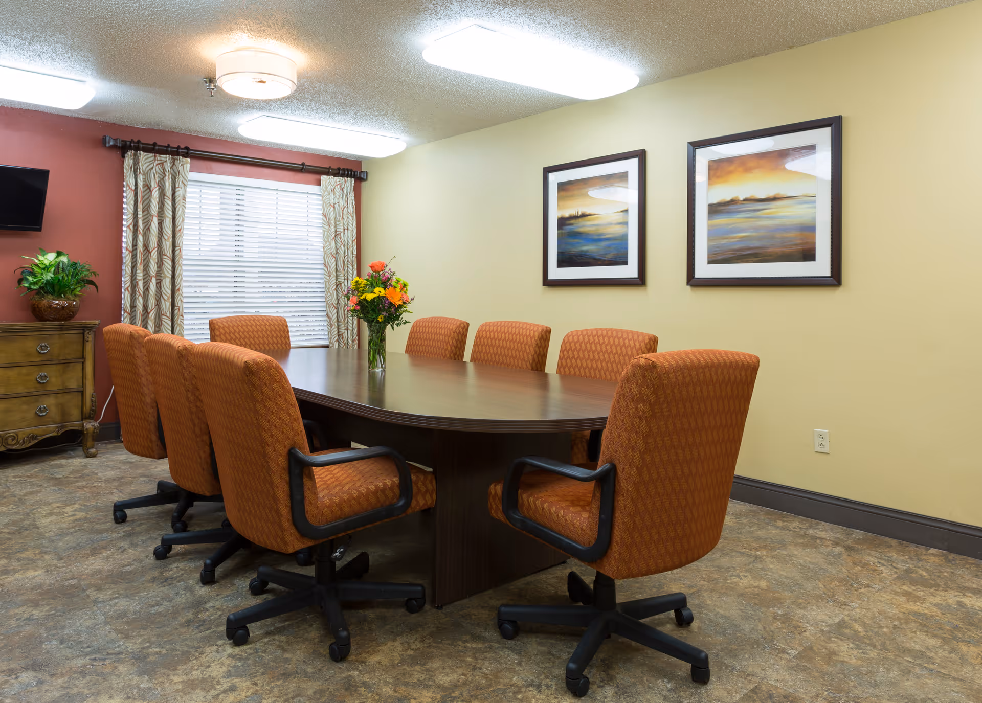Conference-style dining room with a long wooden table surrounded by orange upholstered swivel chairs, framed landscape artwork, a window with curtains, and a vase of flowers on the table.