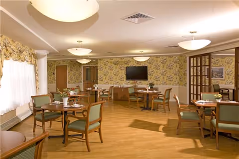 Dining room with round wooden tables, green-upholstered chairs, floral wallpaper, ceiling lights and a wall-mounted TV.