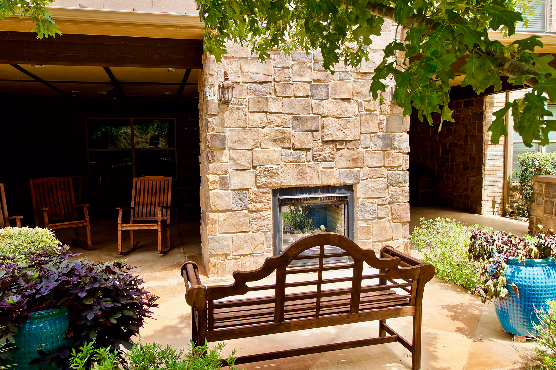 Outdoor seating area with a wooden bench facing a stone fireplace. There are two wooden chairs in a shaded area behind the fireplace, surrounded by green plants and trees.