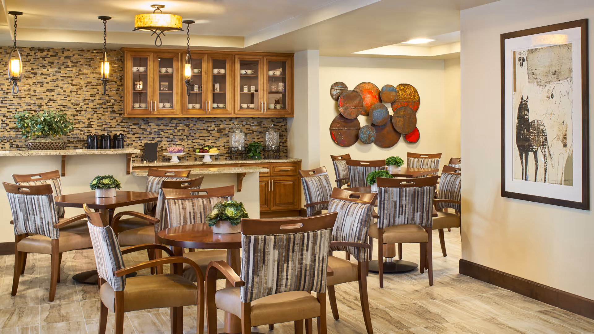 A cozy dining area with several wooden tables and cushioned chairs featuring striped upholstery. The back wall has a mosaic tile backsplash with wooden cabinets above a granite countertop. Decorative plants, fruit bowls, and glass containers are placed on the counter. Warm pendant lights hang from the ceiling, and the walls are adorned with colorful circular metal wall art and a framed abstract painting.