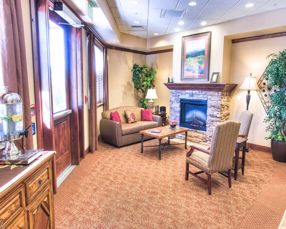 A cozy living room area in a memory care residence featuring a stone fireplace with a framed picture above it, a brown sofa with red and patterned cushions, two striped armchairs, a wooden coffee table, potted plants, and a water dispenser on a wooden cabinet near the entrance door.