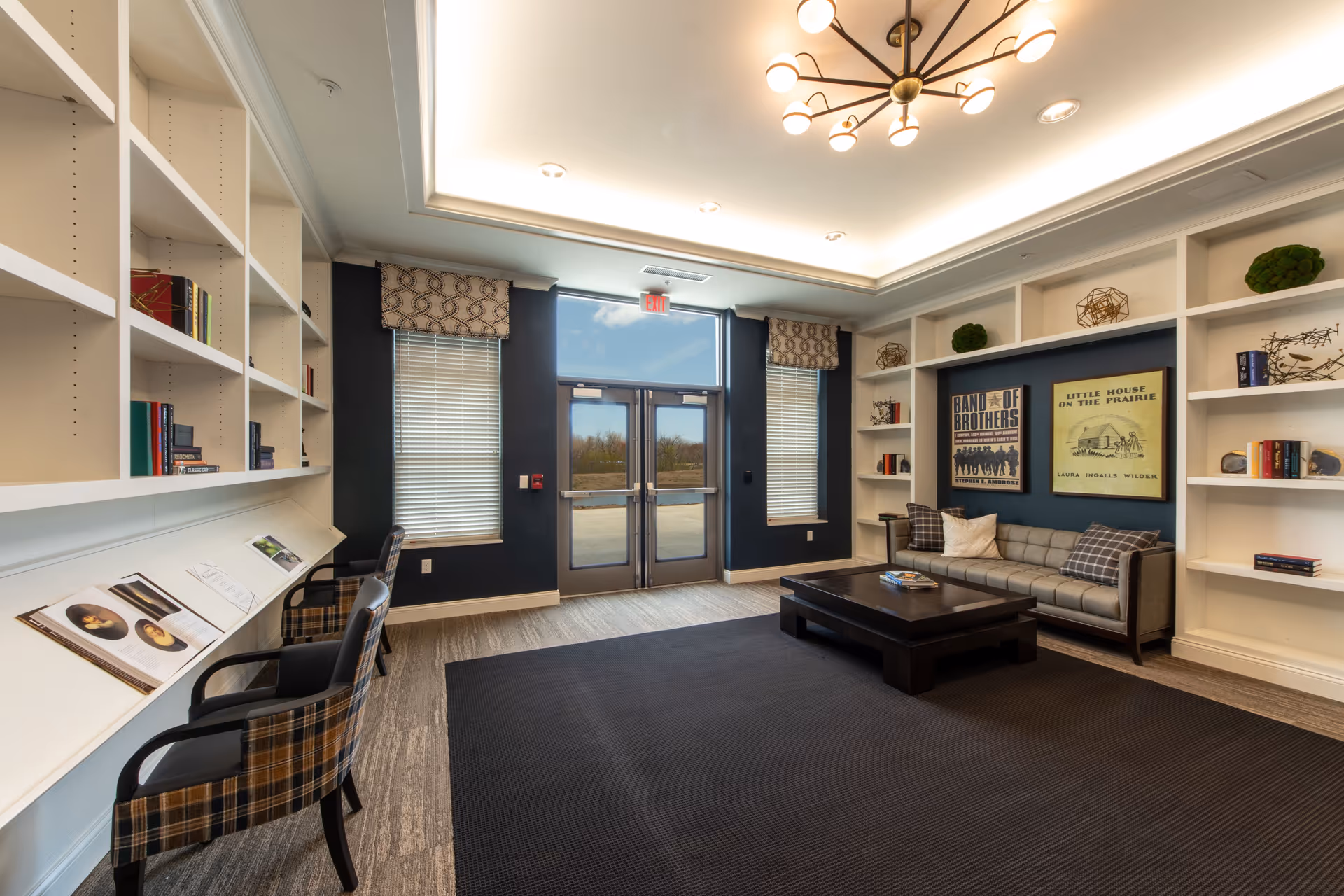 A cozy lounge area with a gray sofa adorned with pillows, a dark wooden coffee table, and built-in white bookshelves filled with books and decorative items. Two large framed posters hang on a dark blue accent wall behind the sofa. Opposite the sofa, there are two plaid armchairs and a long white desk with open books displayed. The room has large windows with blinds and patterned valances, and a glass double door leading outside. The ceiling features recessed lighting and a modern chandelier.