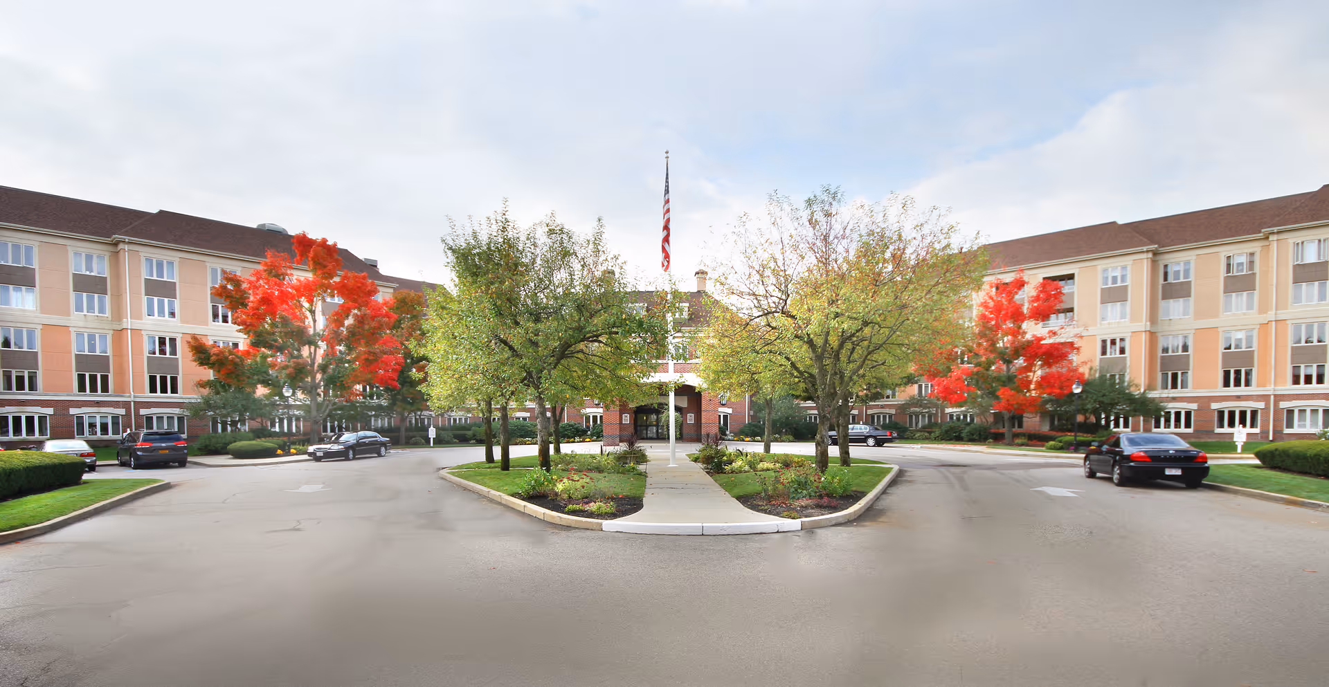 Front exterior view of Fox Hill Village senior living facility with a circular driveway, landscaped greenery, trees with autumn foliage, and an American flag in the center.